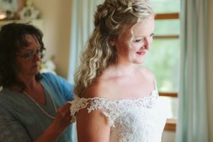 An elegant off-shoulder lace wedding dress being fitted in a sunlit room.