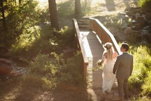 A couple walking along a sunlit wooden bridge surrounded by trees.