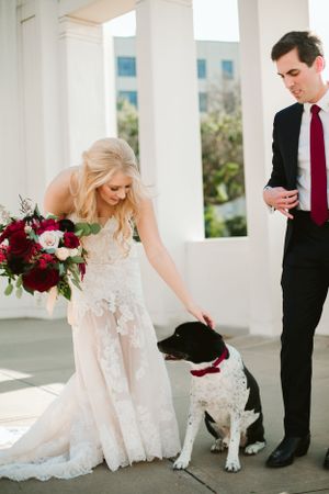 Wedding couple with their black and white dog wearing a red bow tie on a columned porch.