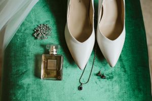 Wedding accessories including white heels, perfume and jewelry on emerald velvet surface.