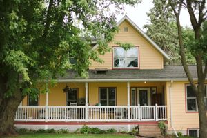 A charming yellow farmhouse with white trim featuring a wraparound porch surrounded by mature trees.