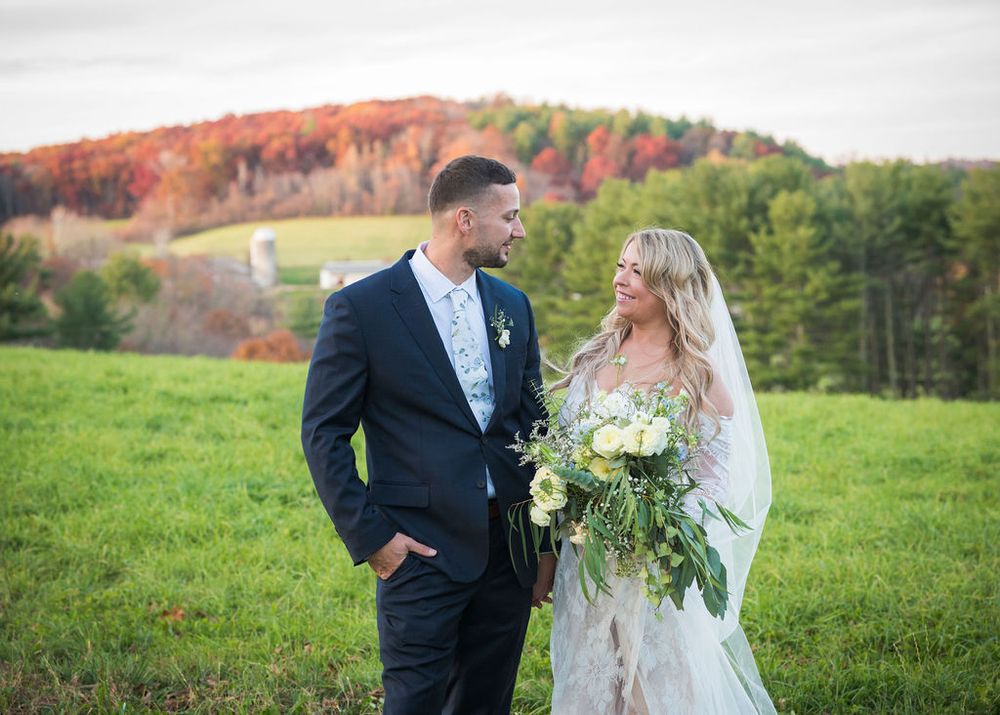 Wedding couple standing in autumn field with colorful fall foliage on hillside.