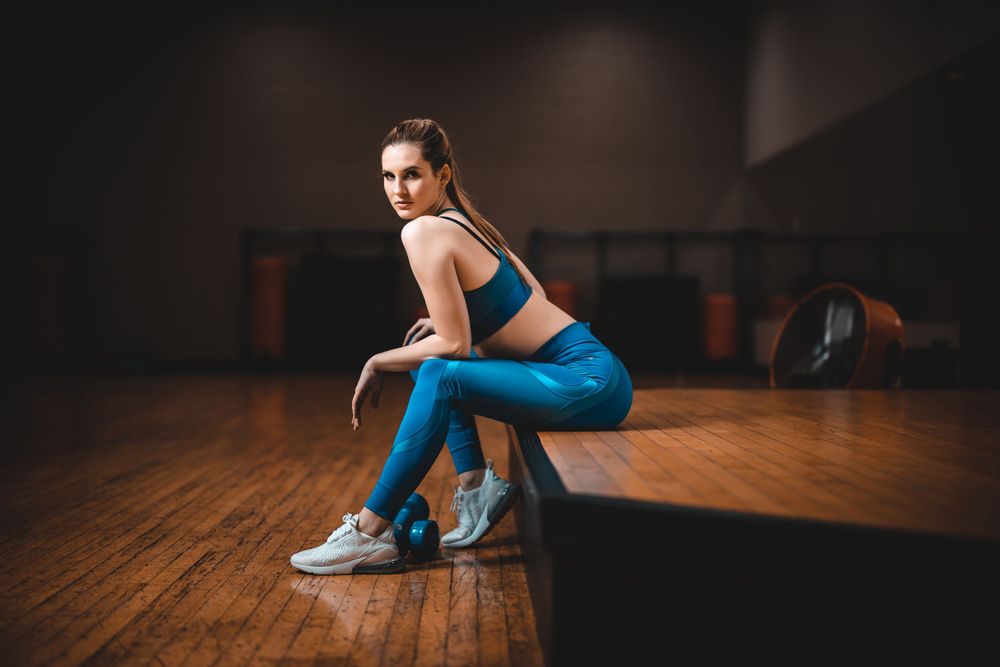 Athletic figure in blue workout gear posing on bench in dimly lit gym setting.