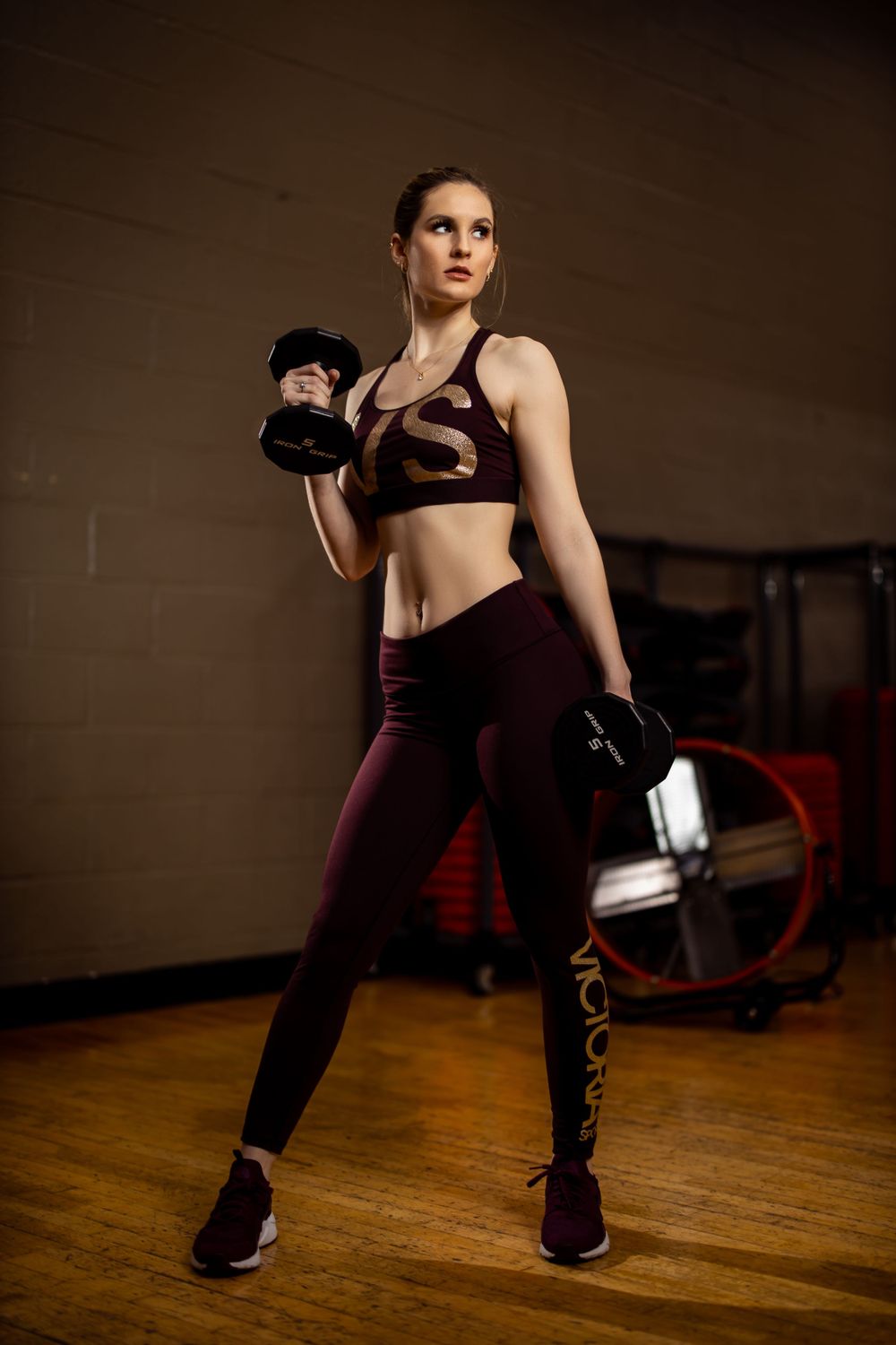 Fitness model working out with dumbbell in burgundy athletic wear at gym.