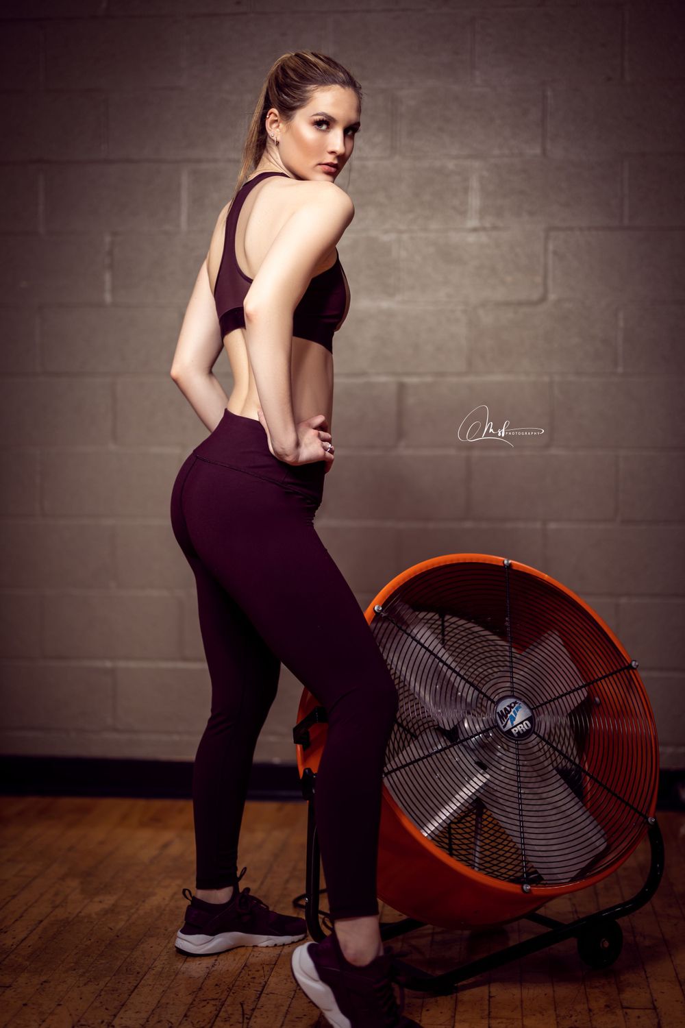 Person in purple athletic wear poses next to large industrial fan in gym setting.