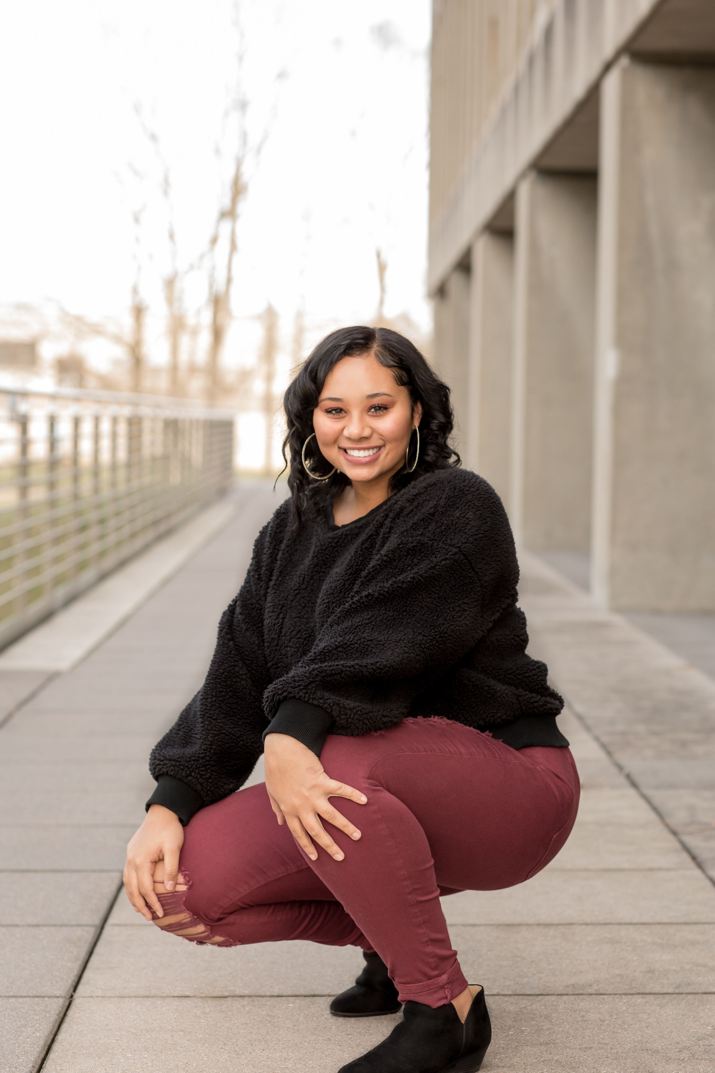 Smiling young woman in black sweater and burgundy pants crouching on an urban walkway.