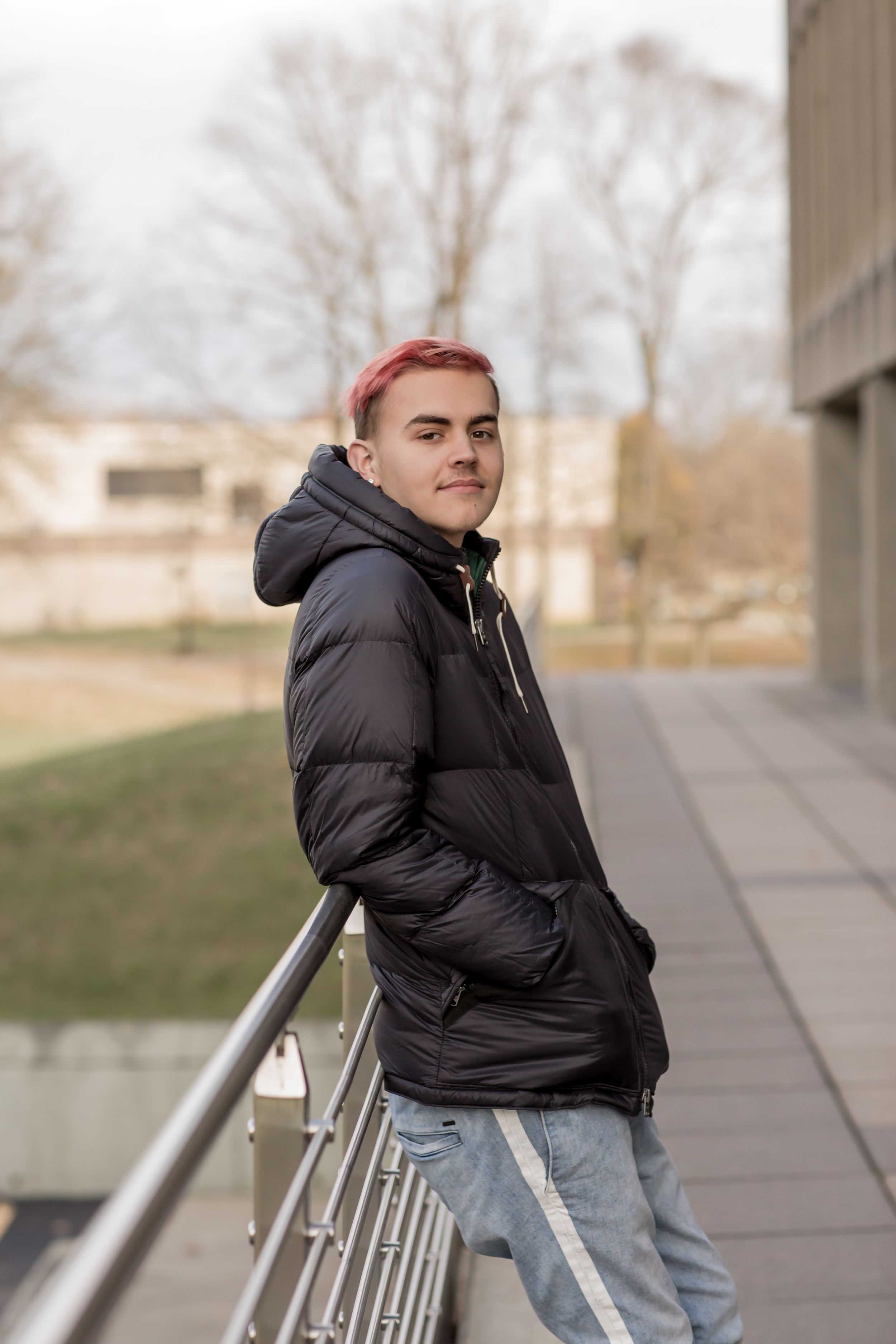 Young person with pink hair leaning on railing in black puffer jacket outdoors.