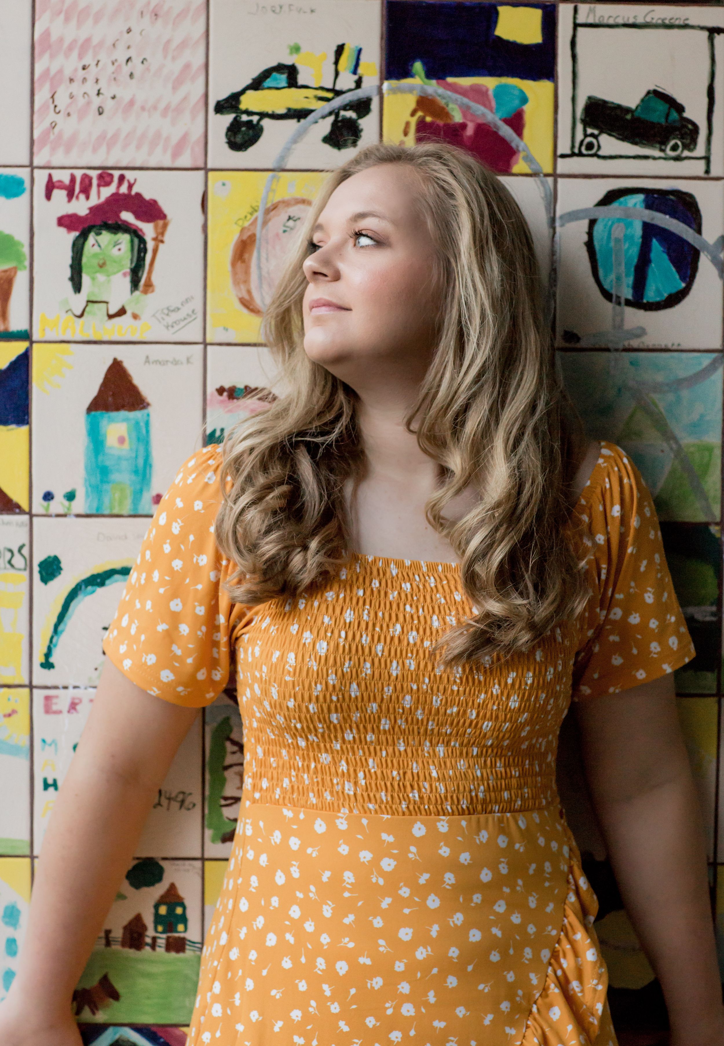 Young woman in orange polka dot dress posing against colorful children's artwork wall.