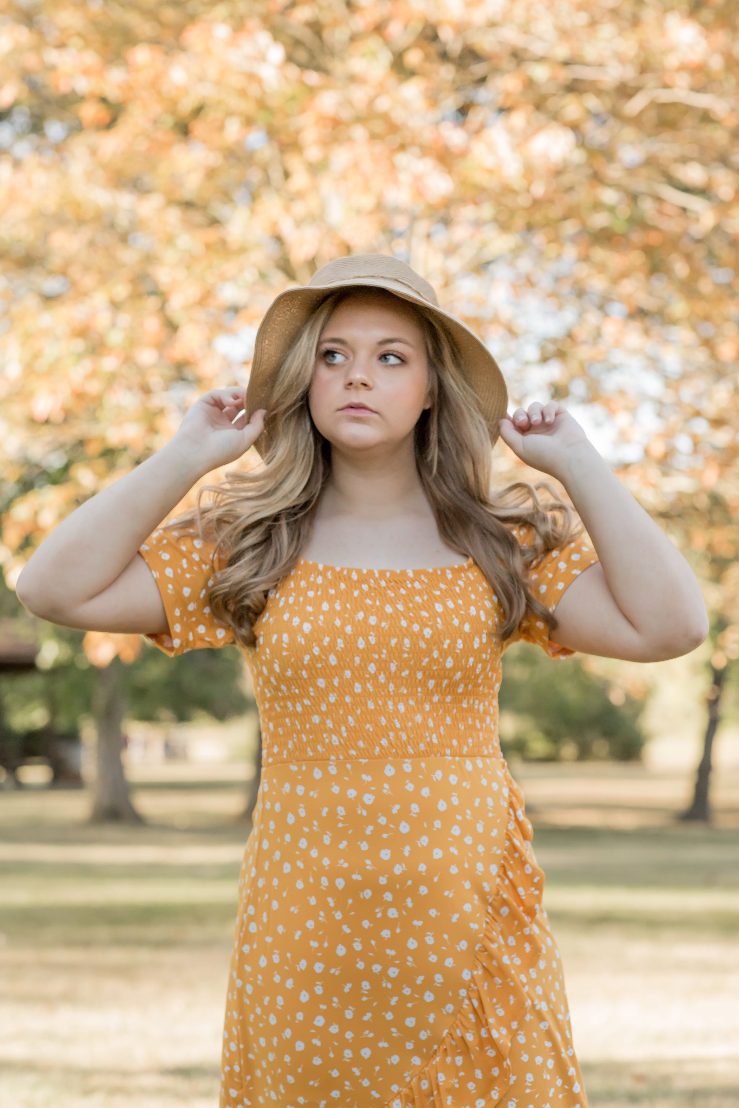 Young woman in orange polka dot dress holding straw hat in autumn park.
