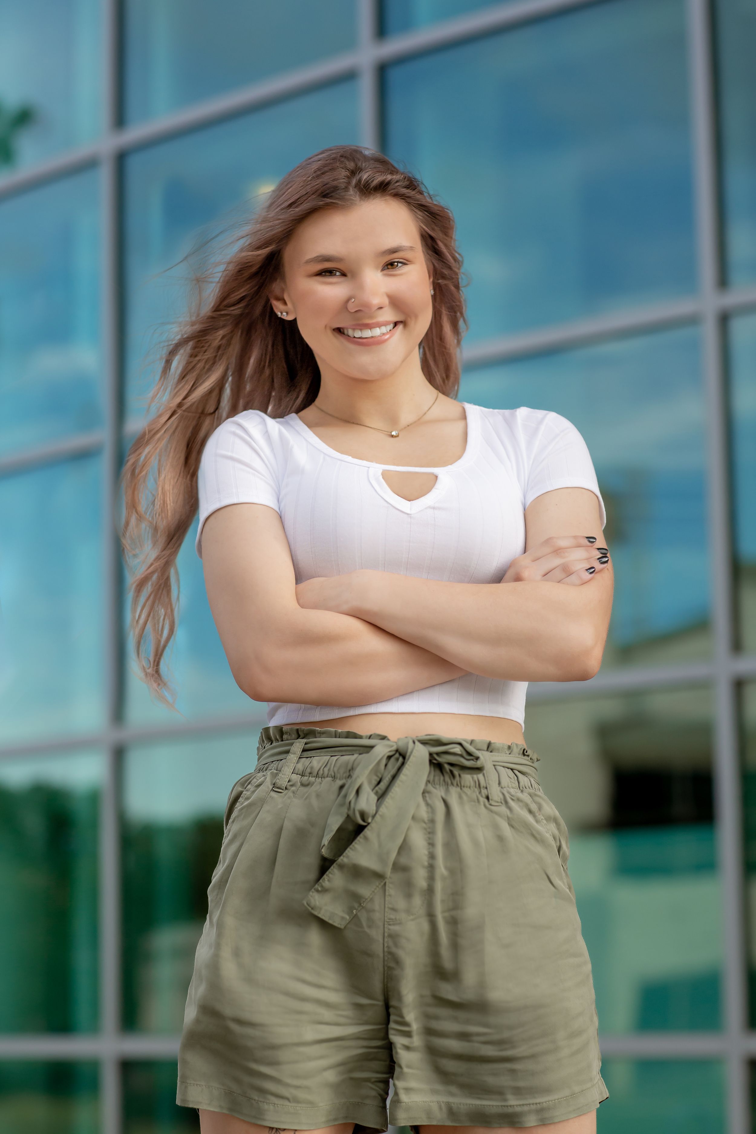 Smiling young woman in white crop top and khaki shorts standing arms crossed before glass building.