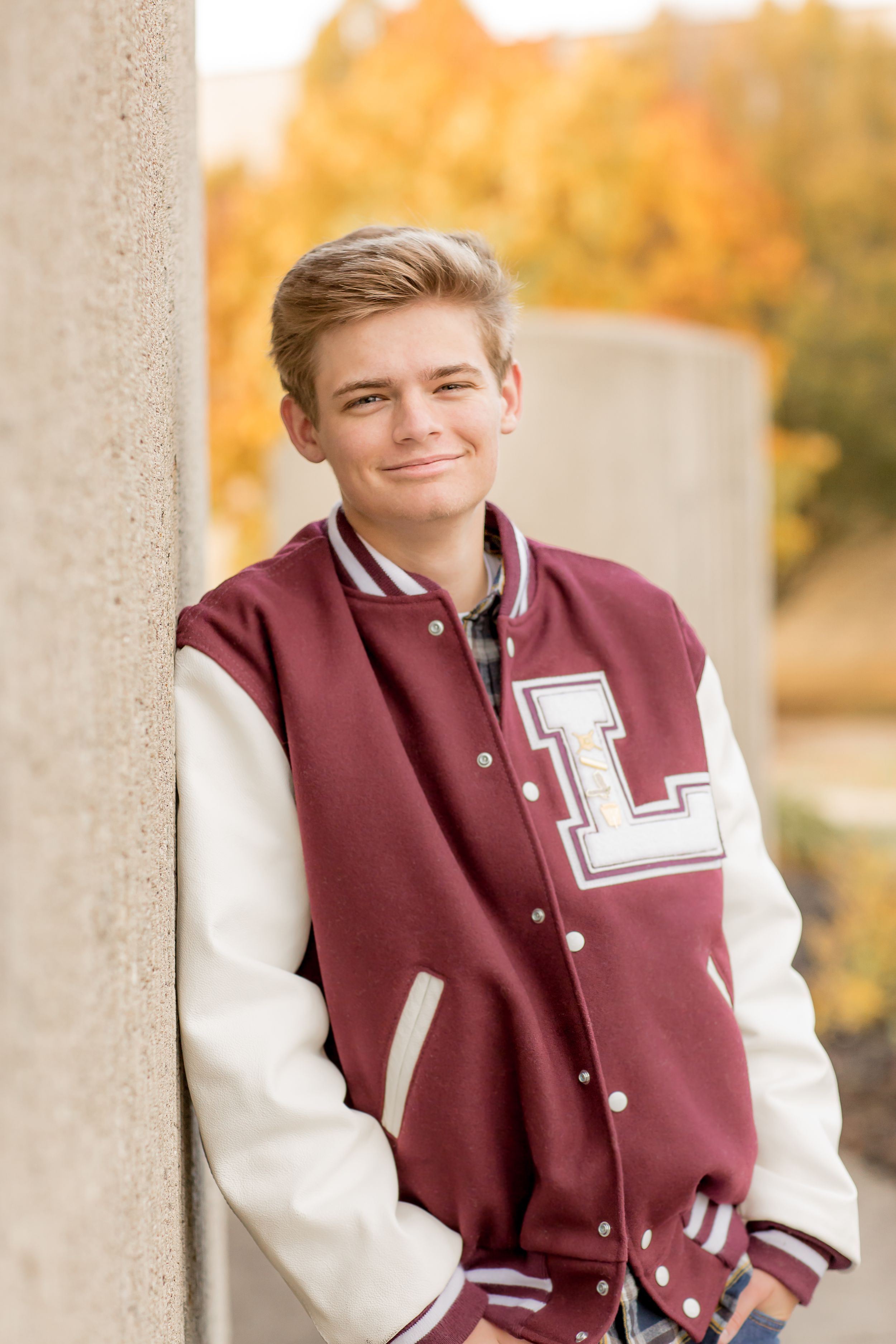 Teen boy in maroon varsity letterman jacket leaning against concrete pillar with autumn foliage background.