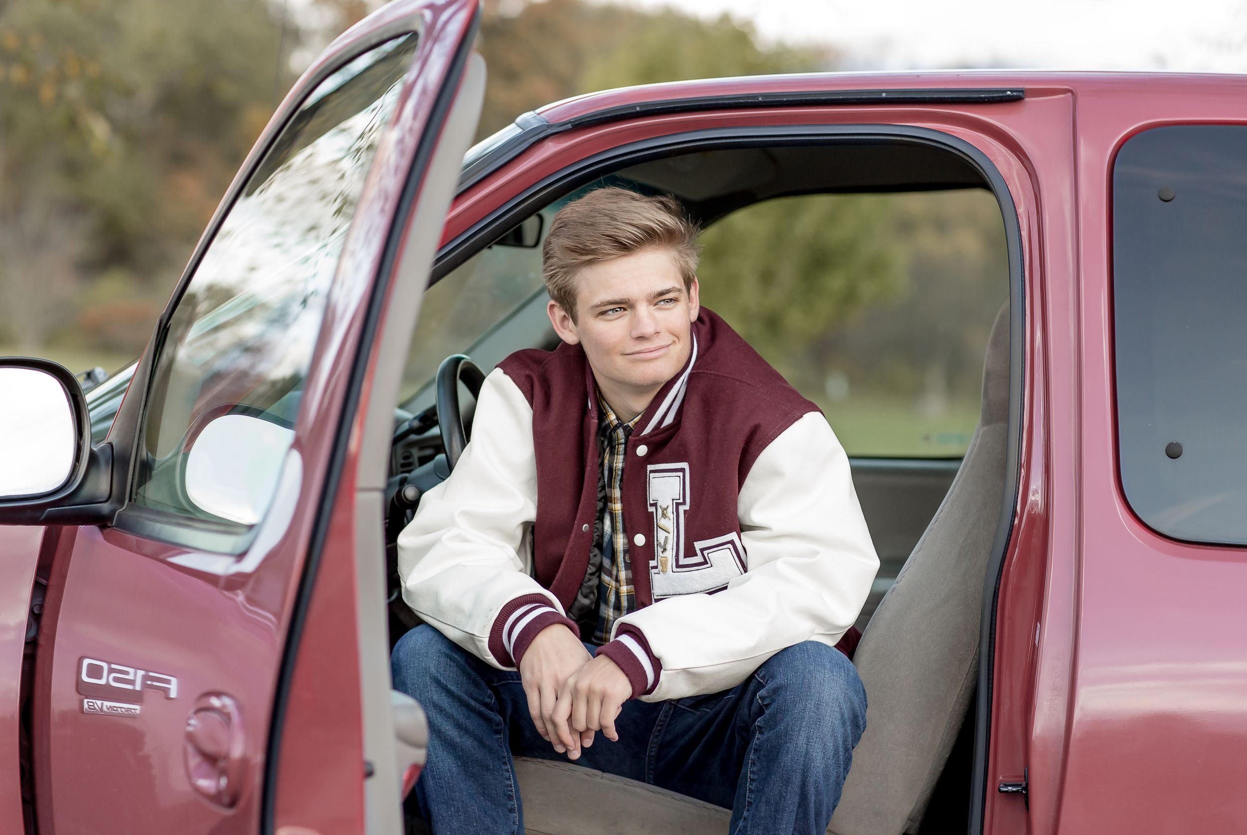Young man in varsity jacket sitting relaxed in open door of red pickup truck outdoors.