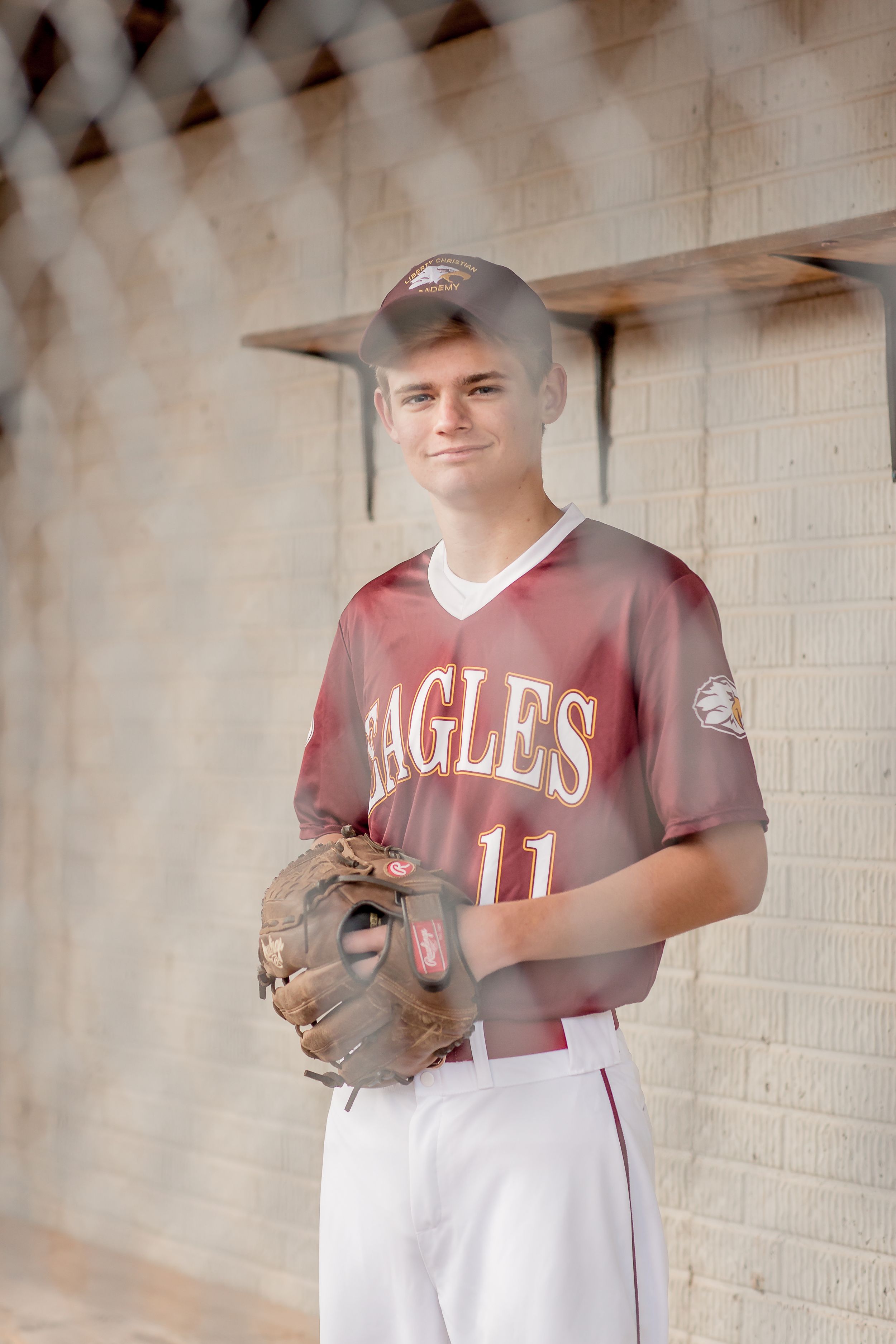 Teen baseball player in Eagles jersey number 11 holding glove in dugout behind chain-link fence.