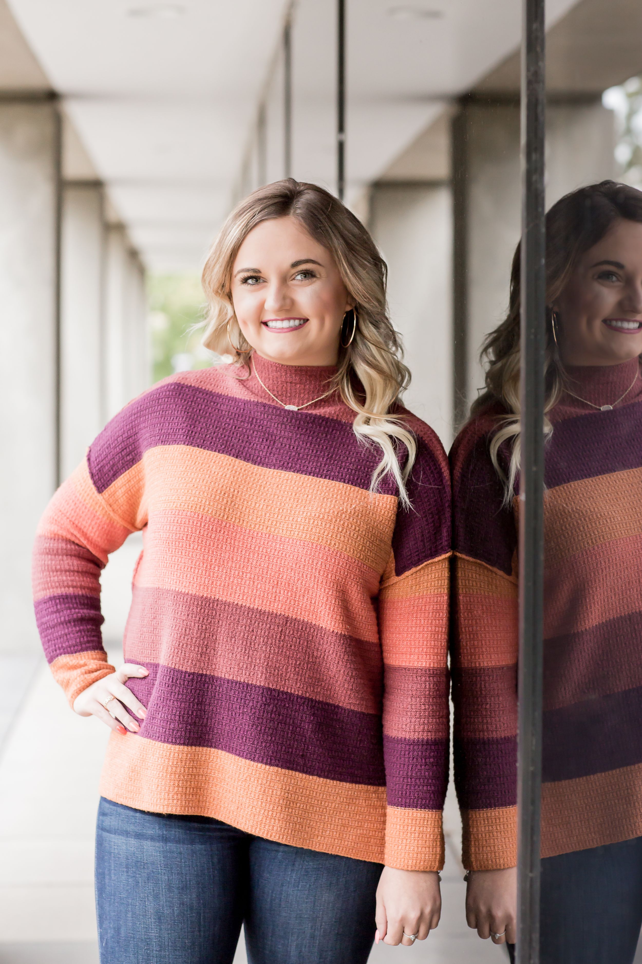 Blonde woman in colorful striped sweater leaning against glass wall with reflection visible.