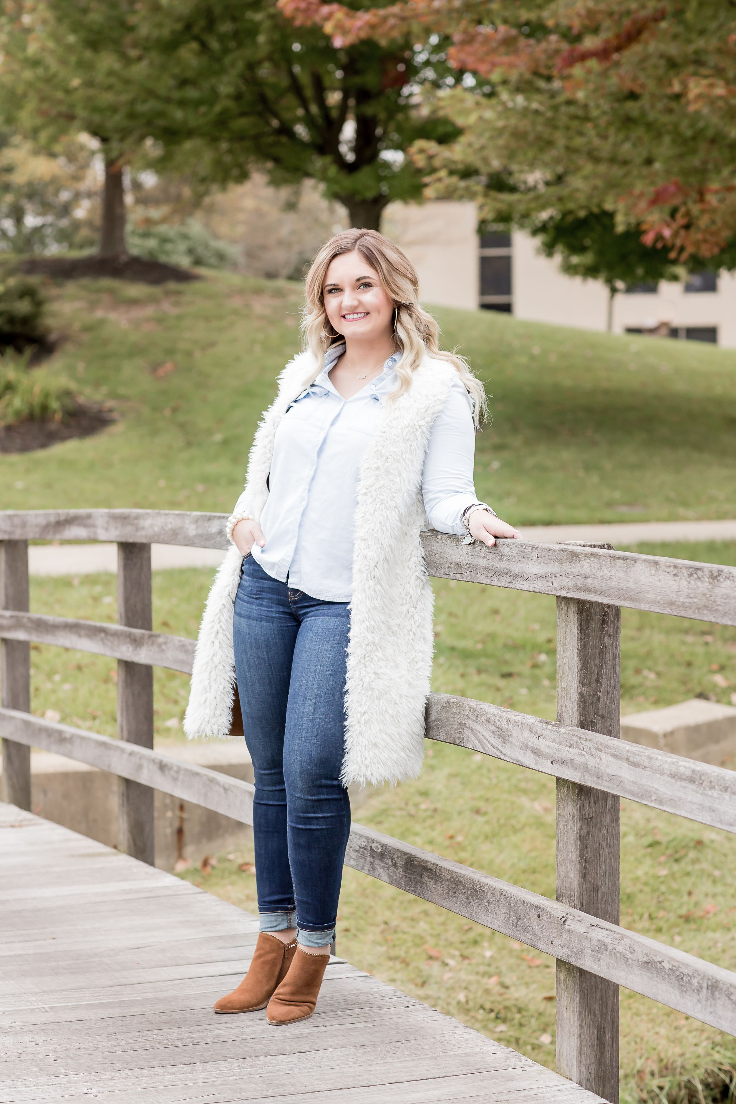 Smiling blonde woman in white fuzzy vest and jeans leaning on a wooden fence outdoors.