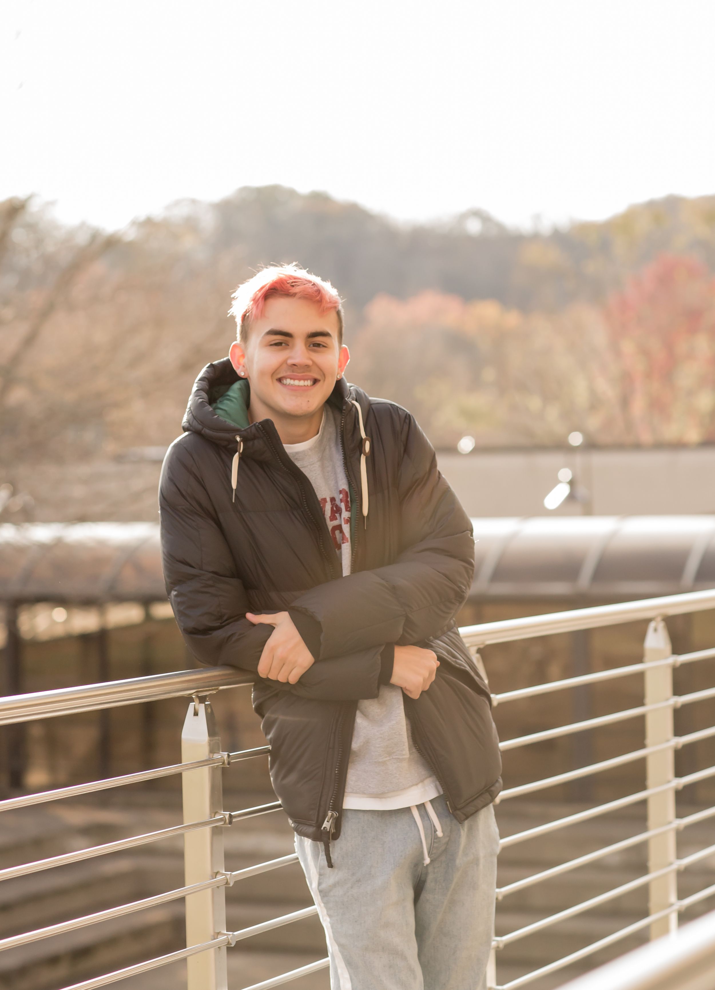 Smiling young person with pink hair leaning on bridge railing in black jacket.