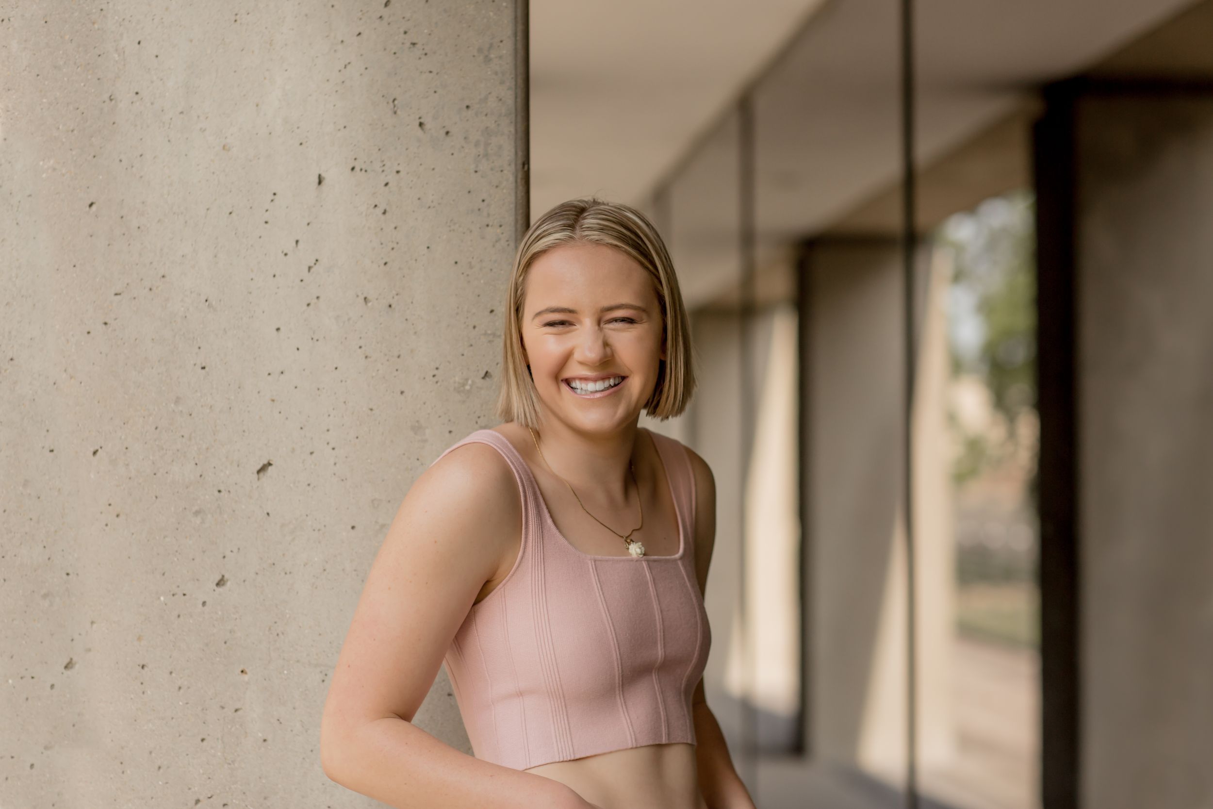 Smiling young blonde woman in pink crop top leaning against concrete column outdoors.