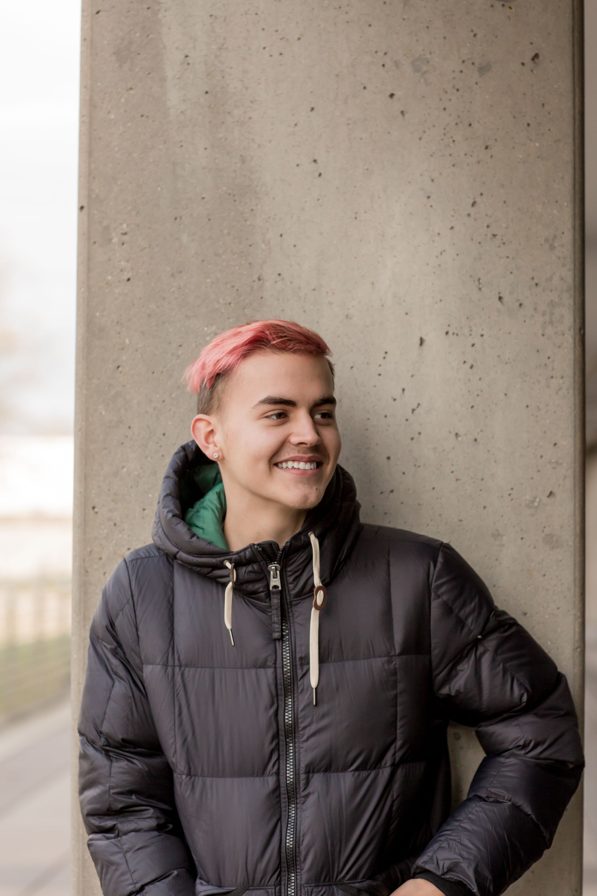 Smiling young person with pink hair leaning against concrete wall in black puffer jacket.