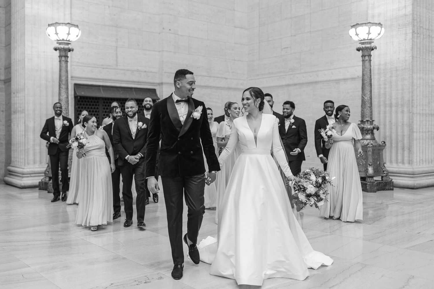 Wedding party walking through a grand stone hallway with elegant lighting and architecture.