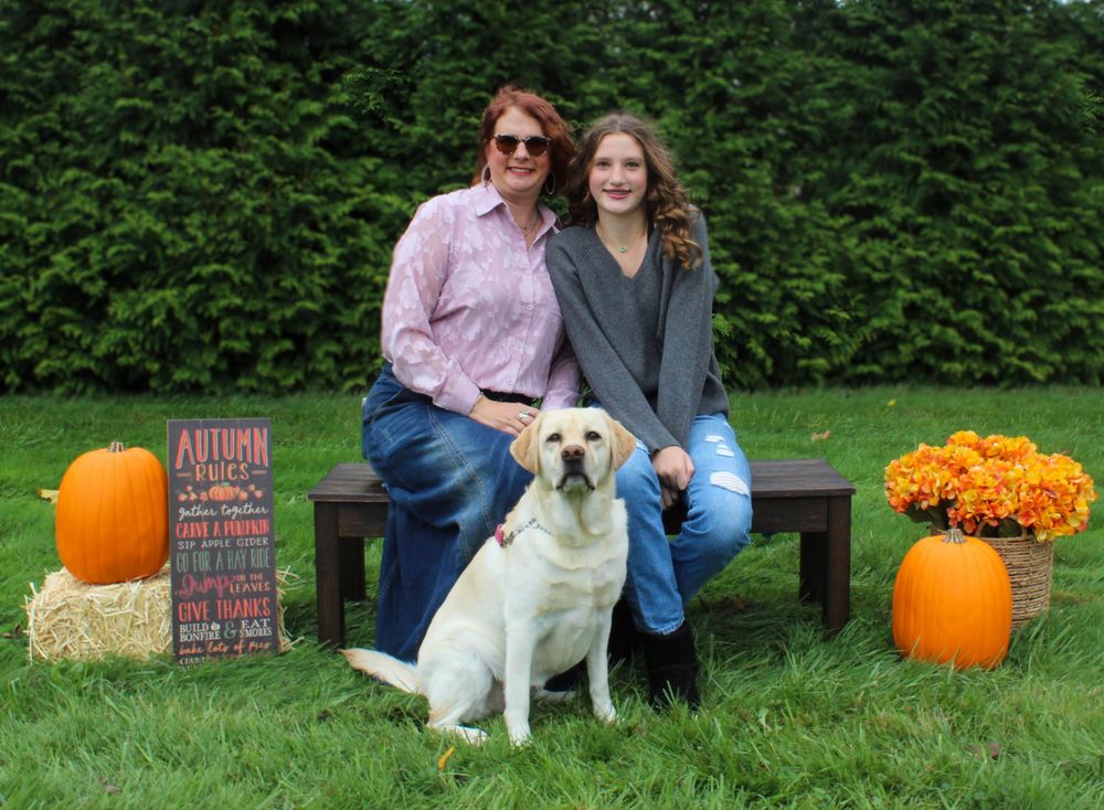 Two people sit with a yellow Labrador on bench surrounded by fall decorations and pumpkins.