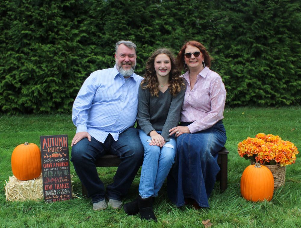 Family poses on wooden bench with autumn decor including orange pumpkins and mums in an outdoor garden setting.