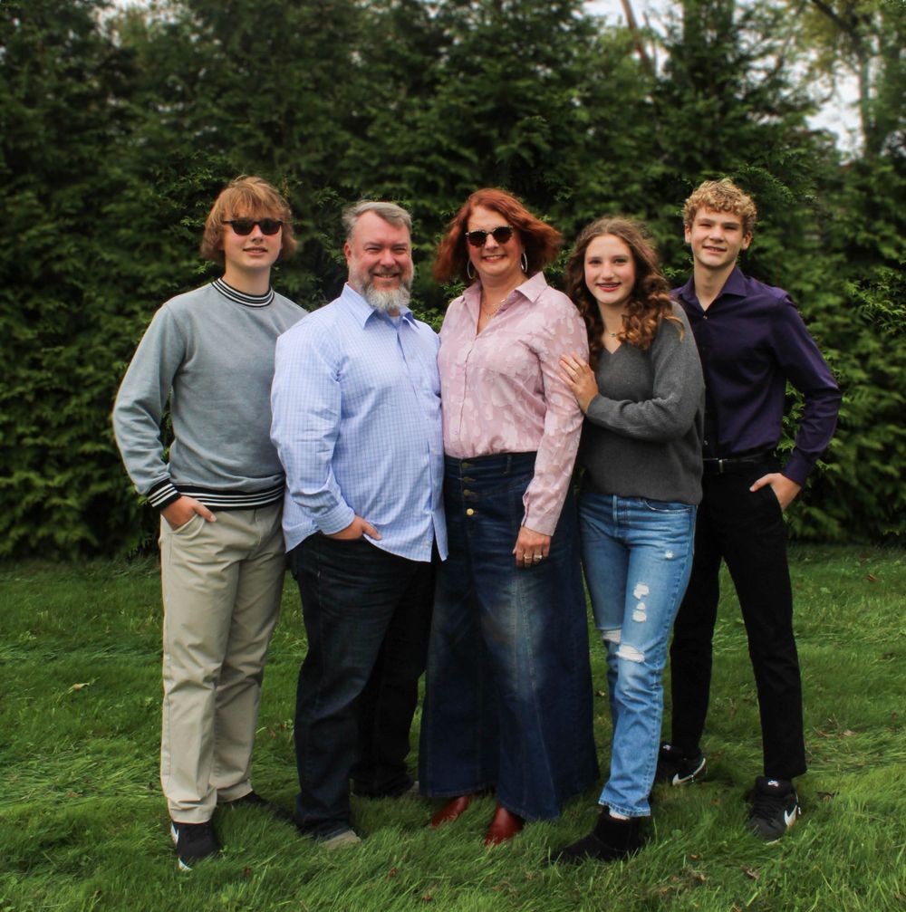 Five people pose together on grass with evergreen hedge in background.
