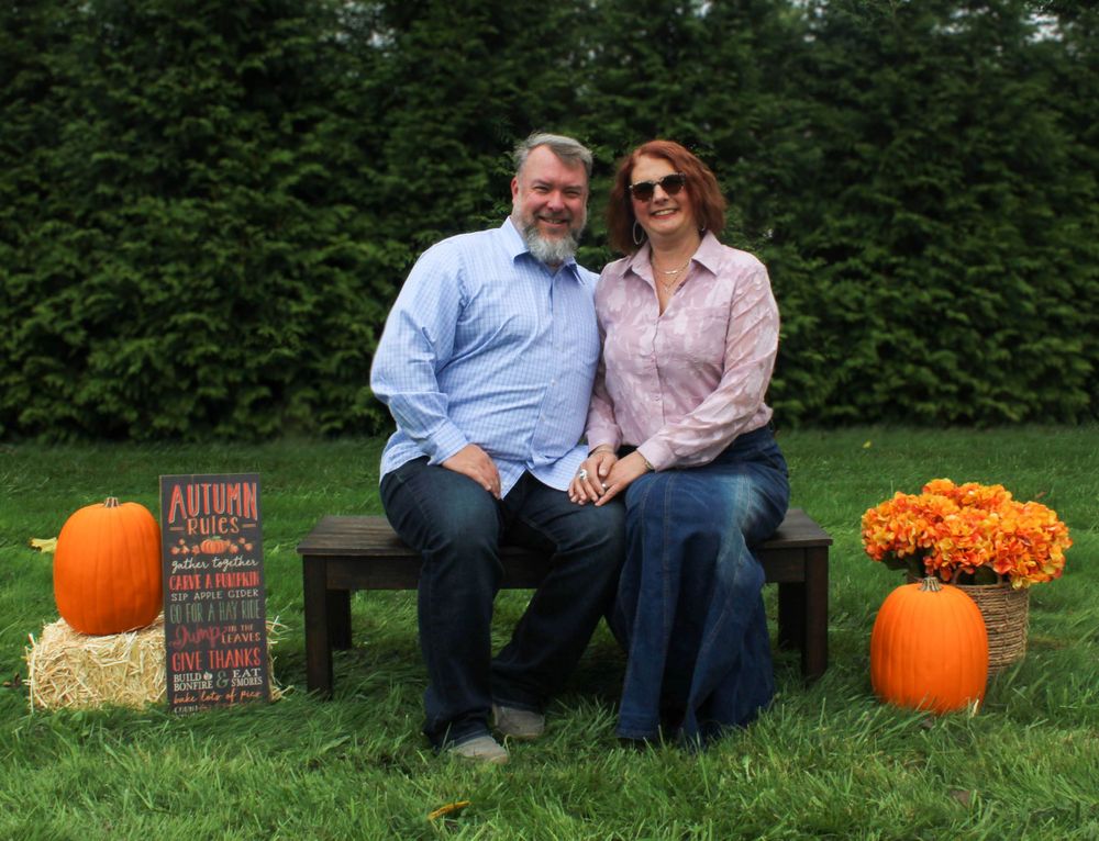 Couple sits on wooden bench with autumn decor including pumpkins and orange mums.