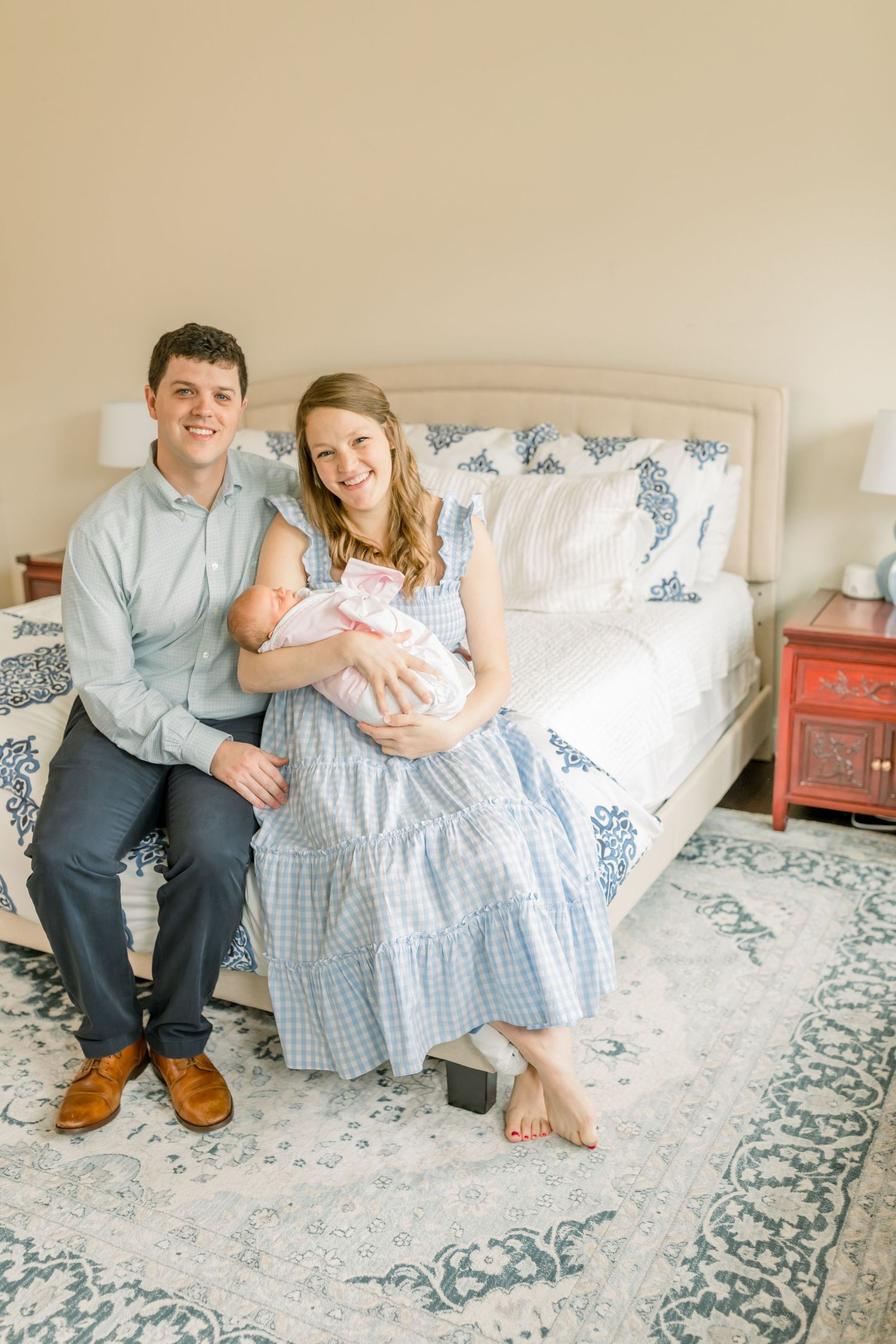 A couple sits on a blue patterned bed with their newborn in a sunny bedroom.