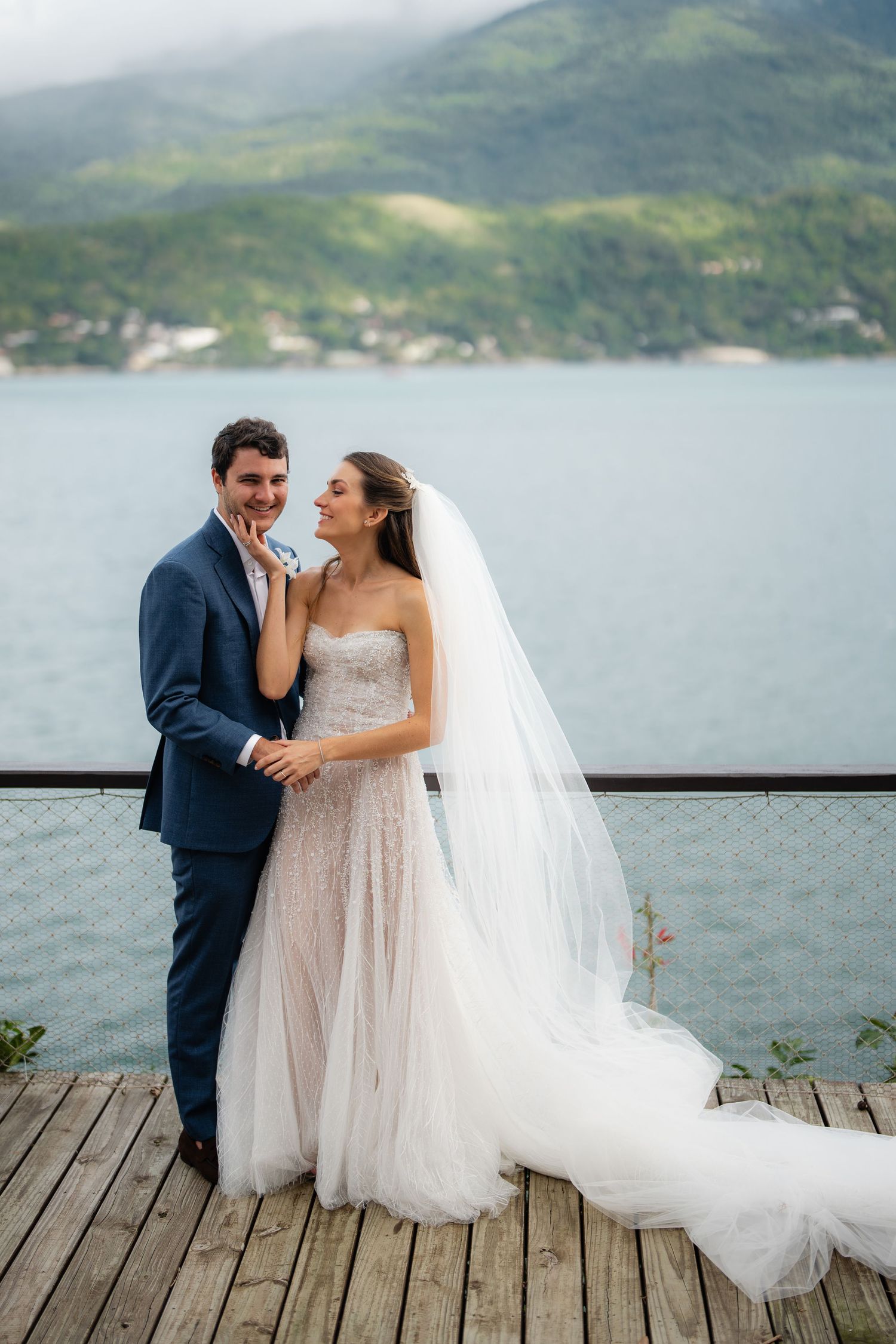 A romantic wedding couple poses on a wooden deck overlooking a scenic mountain lake landscape.