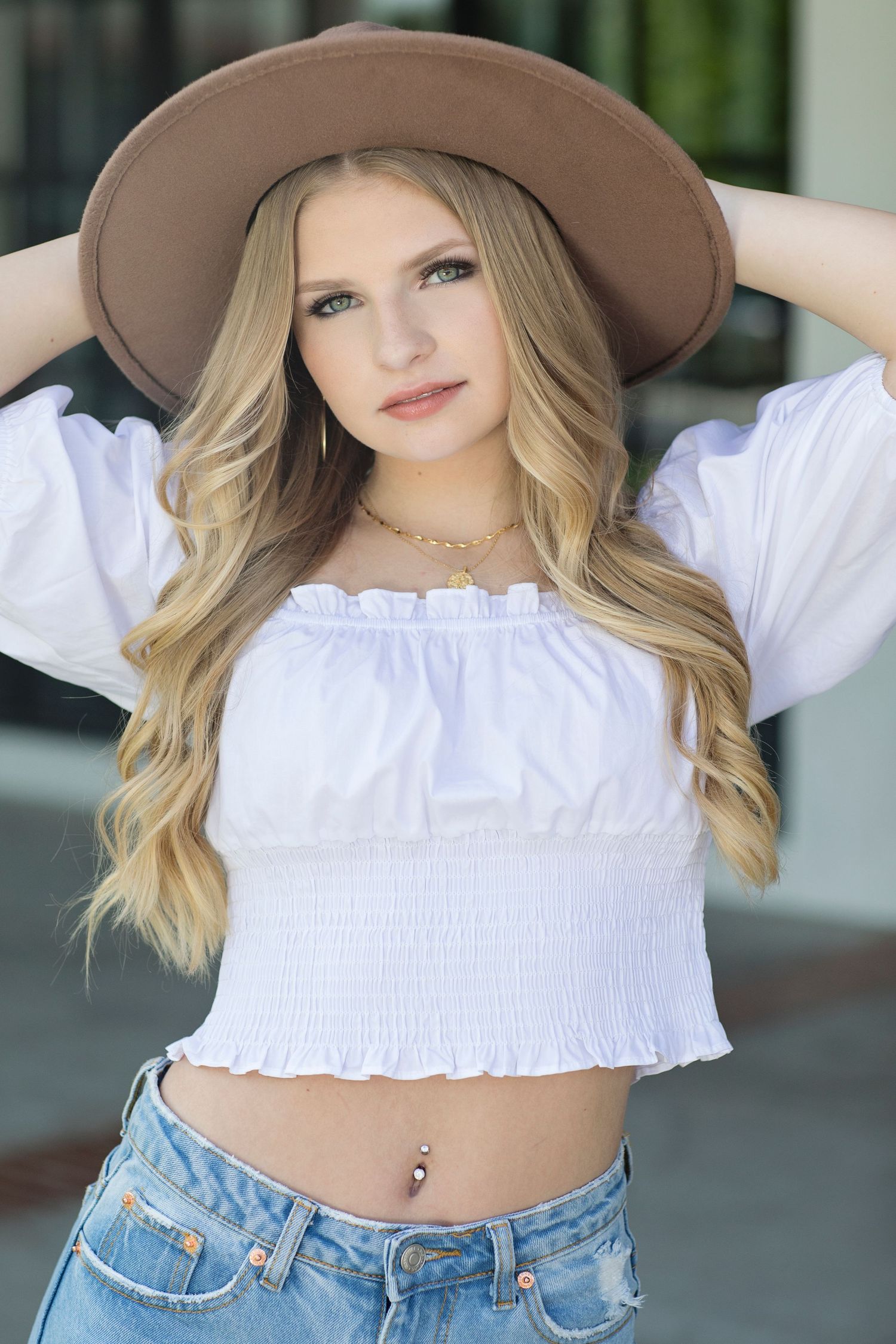 A person in a brown hat and white crop top poses against a blurred background wearing blue jeans.