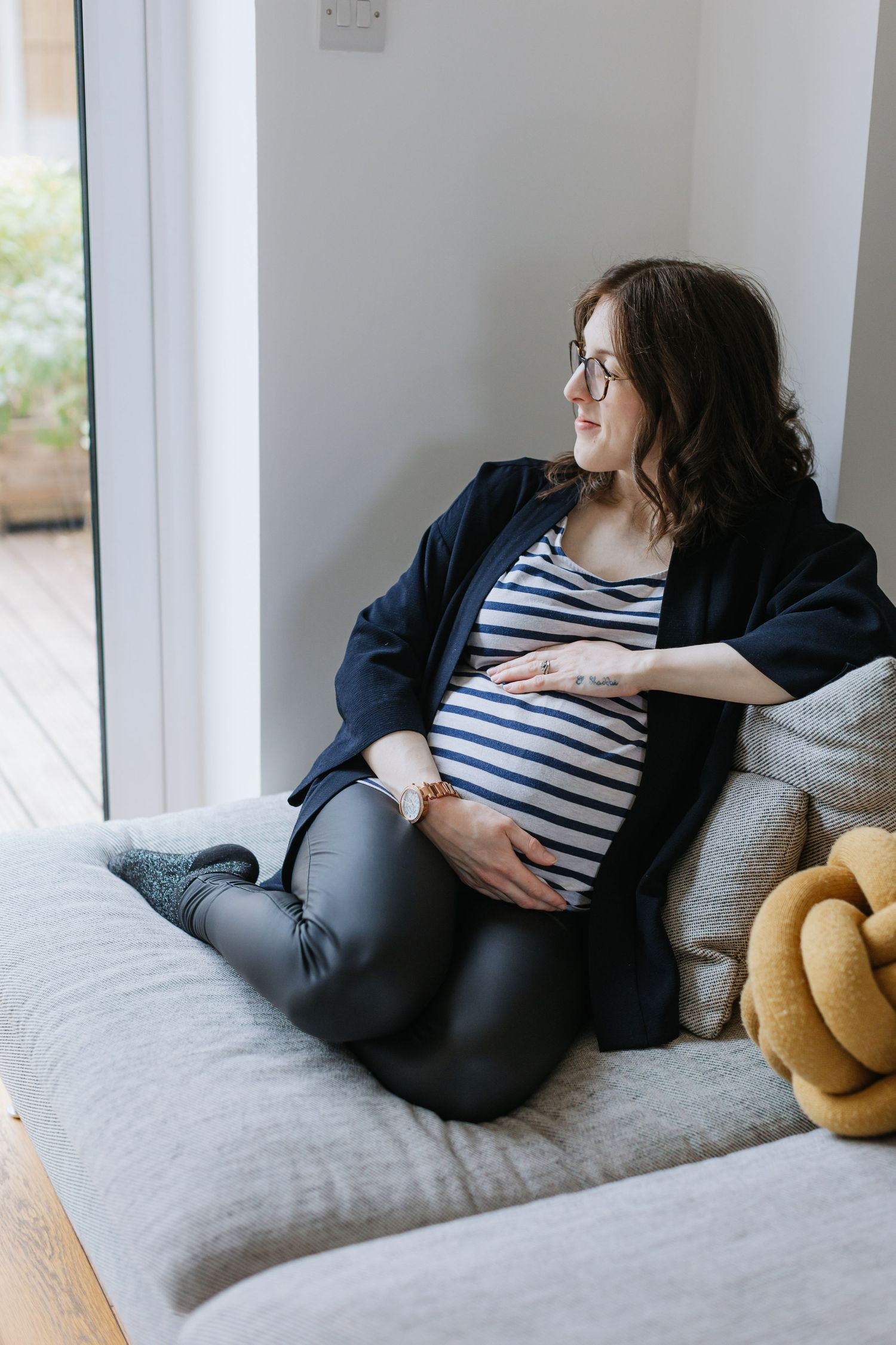 Expectant mother in striped shirt gazes out window while relaxing on grey couch.
