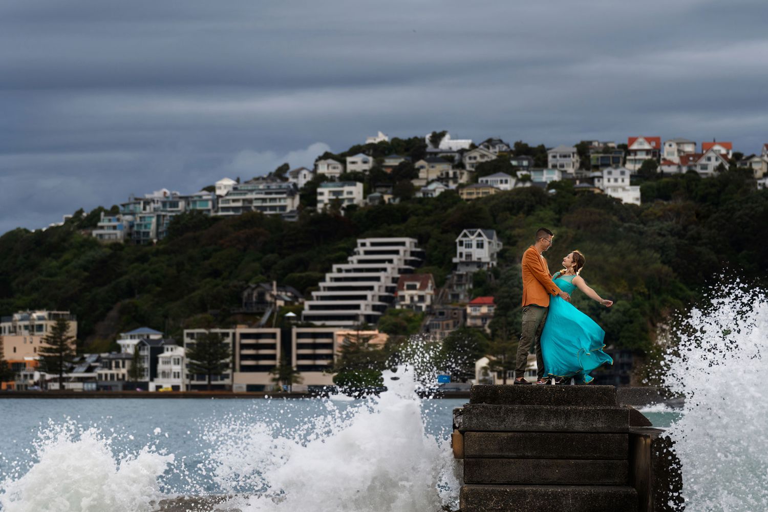 Indian couple holding each other on a rock at Oriental Bay with rising tides and the Wellington city skyline in the background