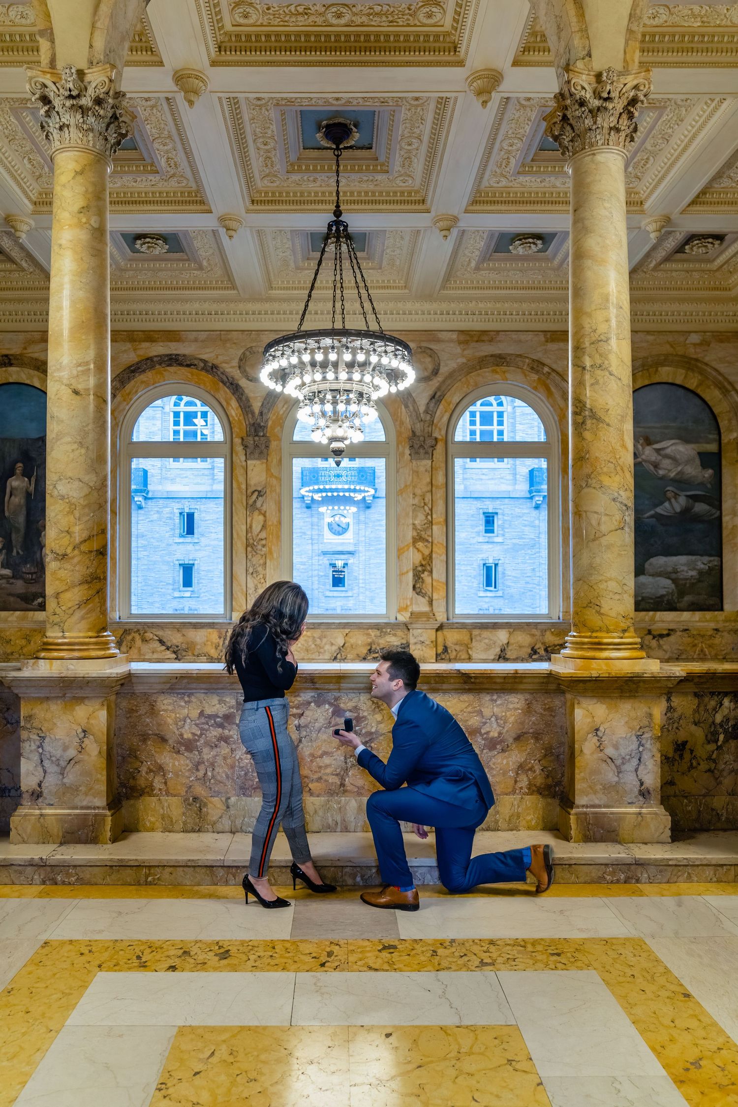 A surprise proposal moment in an elegant marble hall with ornate architecture and chandelier.