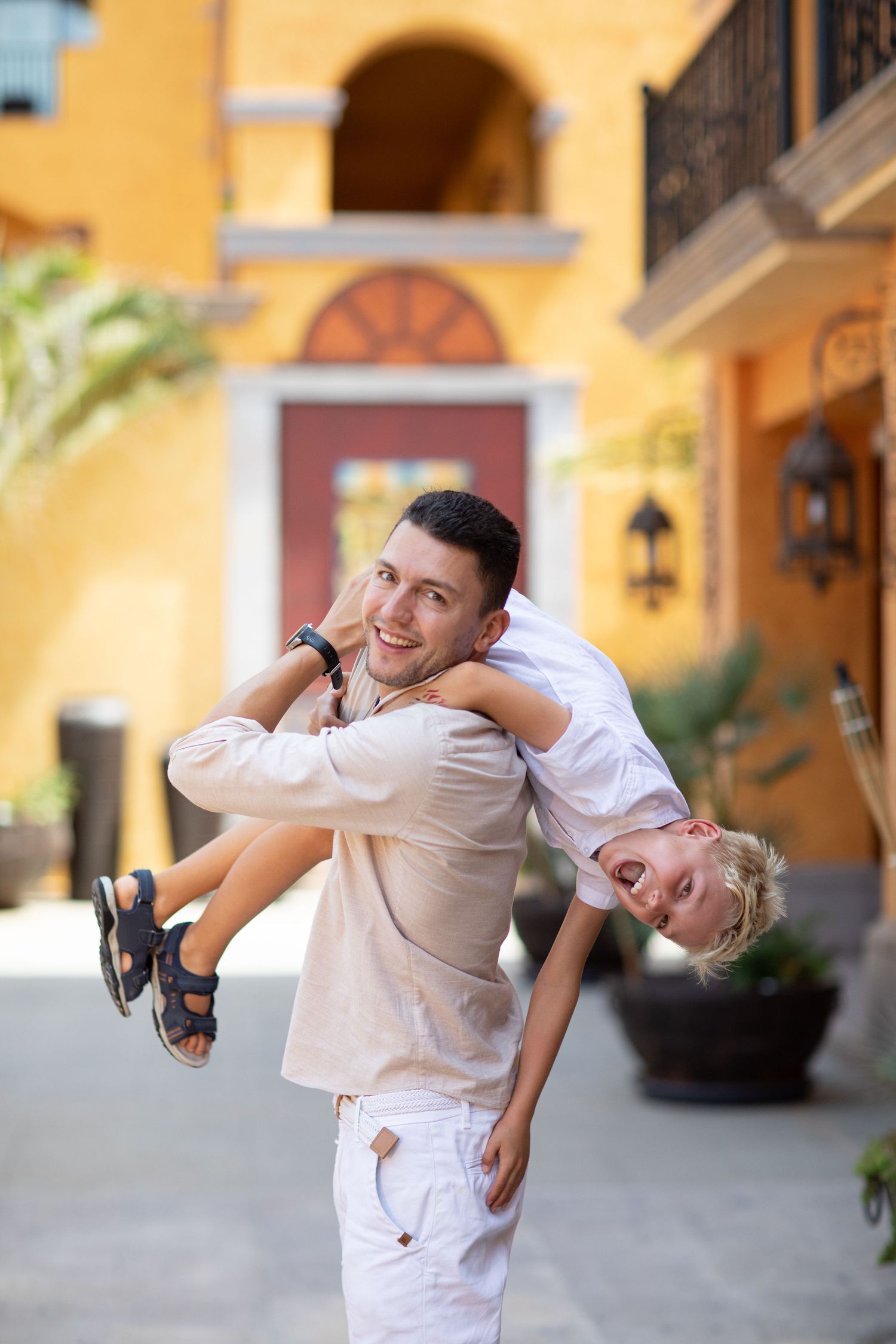 A boy hangs upside down from his dad's back, both smiling, in a sunny courtyard.