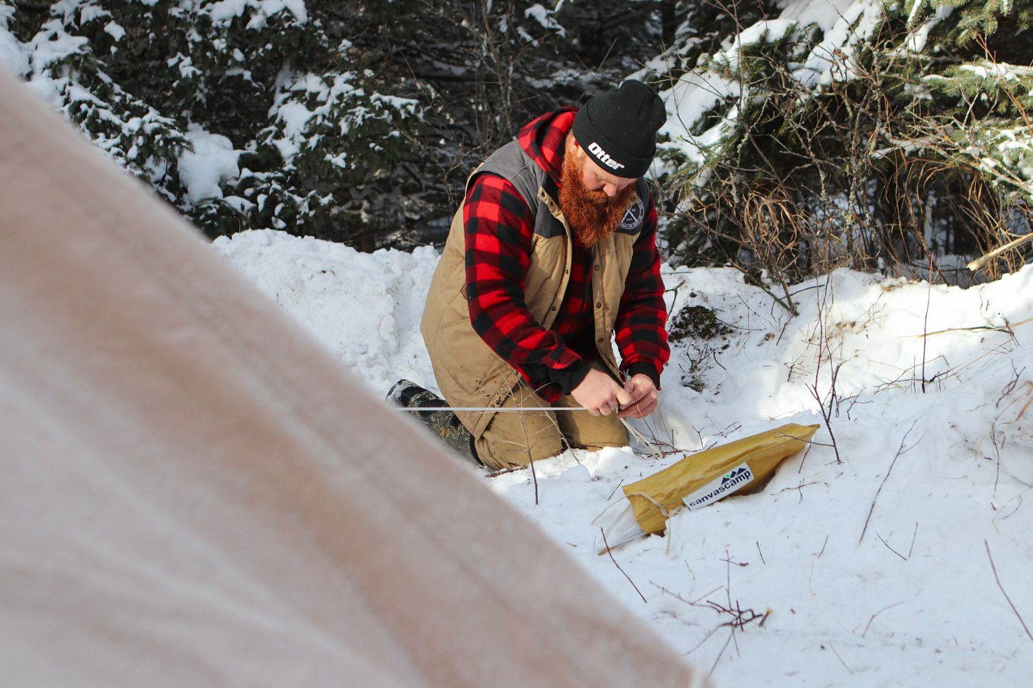 Setting up winter camping gear in snowy forest while wearing red plaid jacket.
