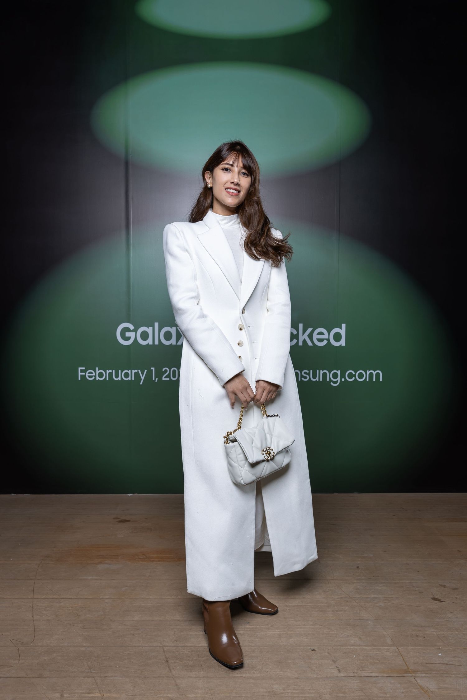 A person in a white coat holding a small handbag against a dark background with spotlights.