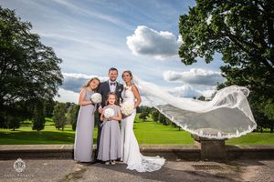 Dramatic photo of small wedding group with flowing veil caught in wind against cloudy sky.