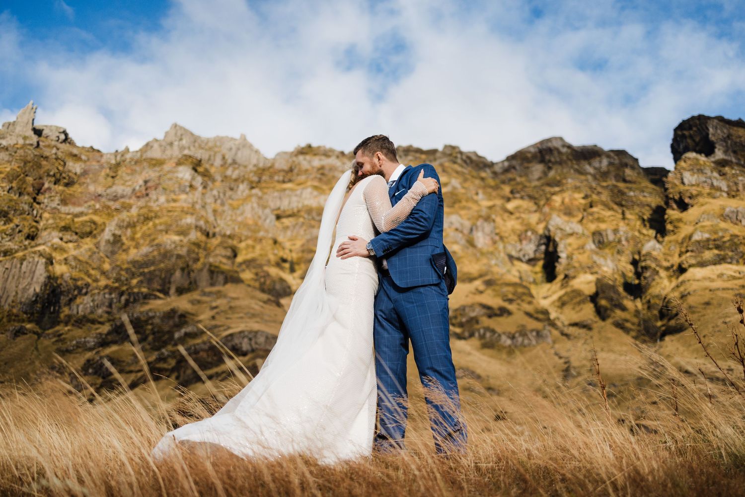 A wedding couple embraces against dramatic rocky cliffs and blue skies.
