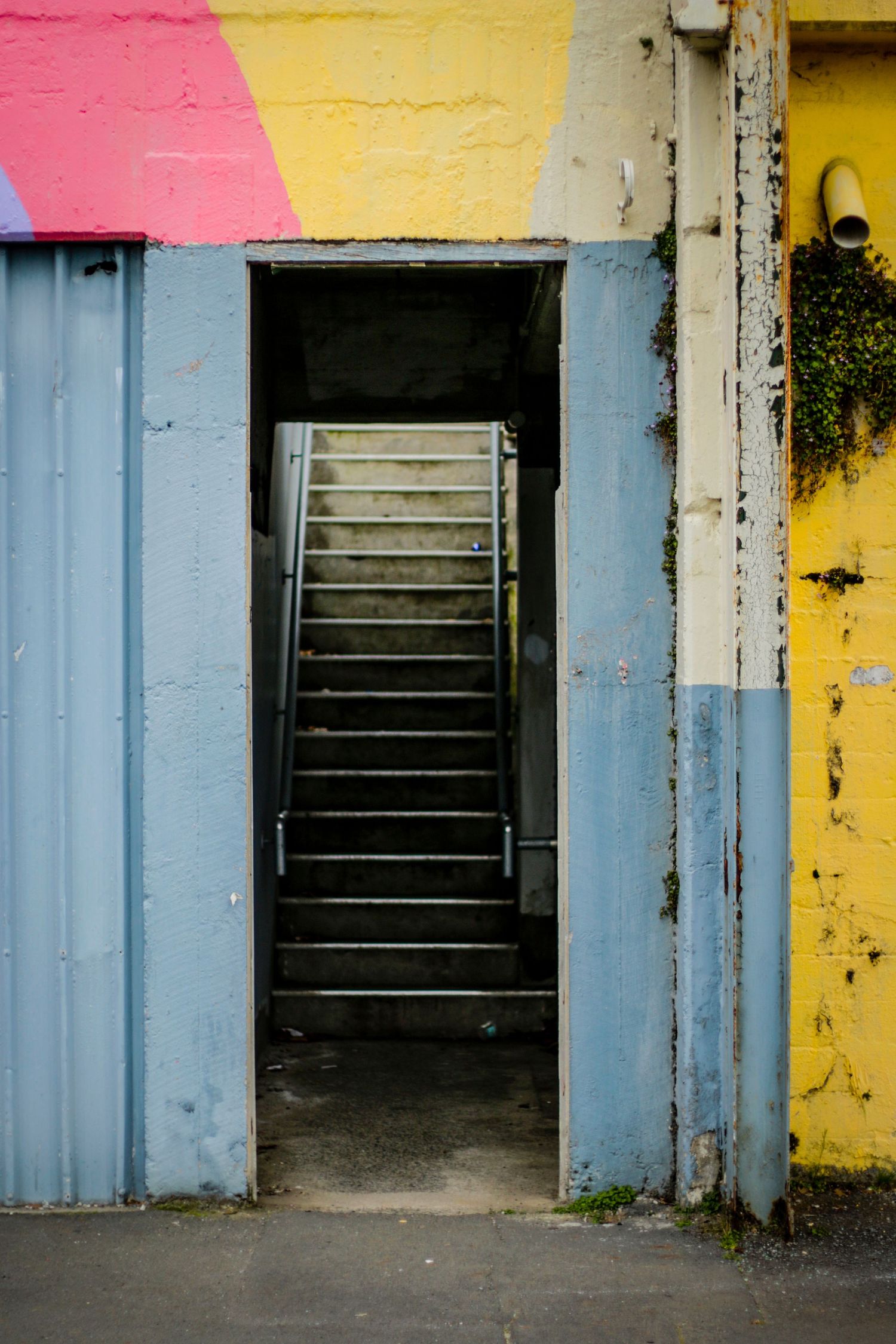 A weathered doorway painted in bright blue, pink, and yellow opens to reveal a dark stairwell leading upward, framed by peeling walls and a