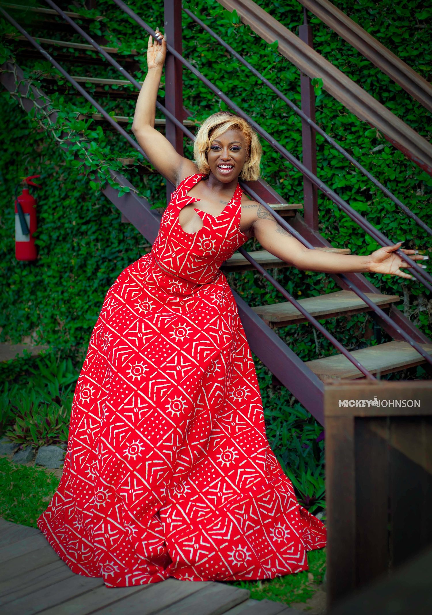 A joyful figure in a flowing red and white patterned dress twirls and dances on a wooden deck surrounded by greenery.