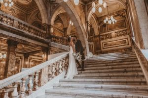 Elegant wedding scene at a grand marble staircase with ornate architecture and chandeliers in warm golden tones.