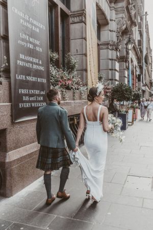 Couple walking hand in hand along historic street, groom in Scottish kilt and bride in sleek white gown.