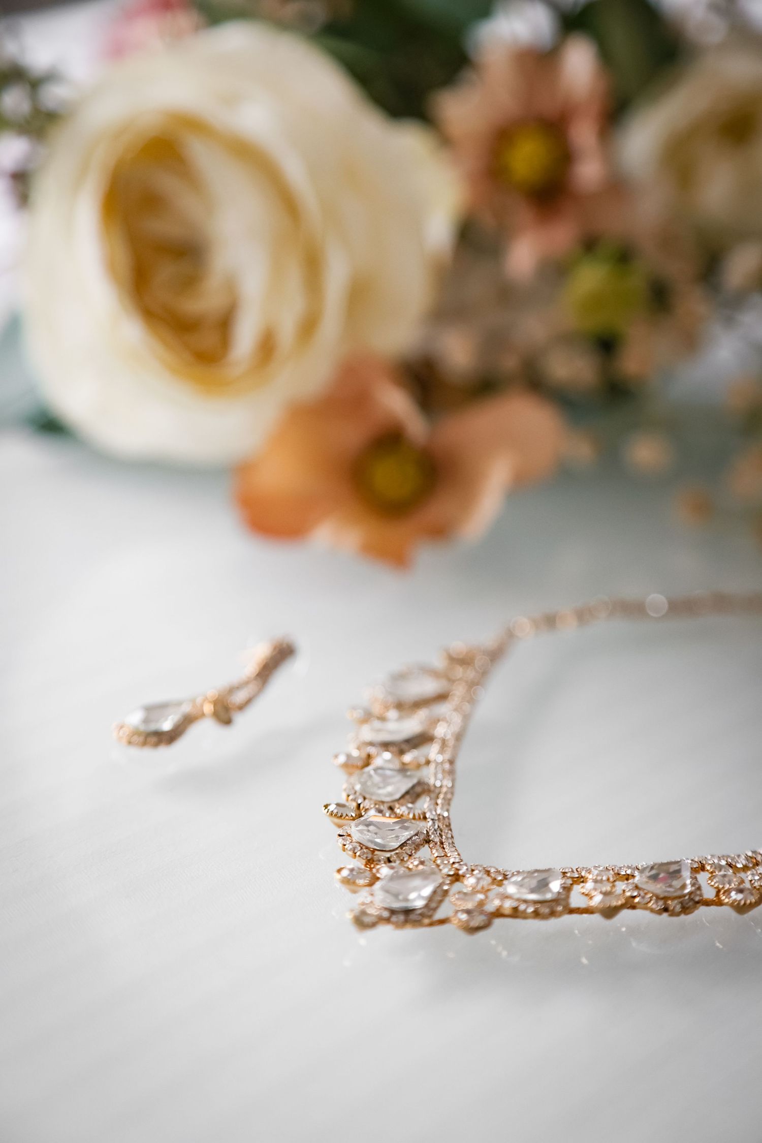 Delicate rose gold bridal jewelry and white roses with peach flowers arranged on white surface in romantic wedding setting.