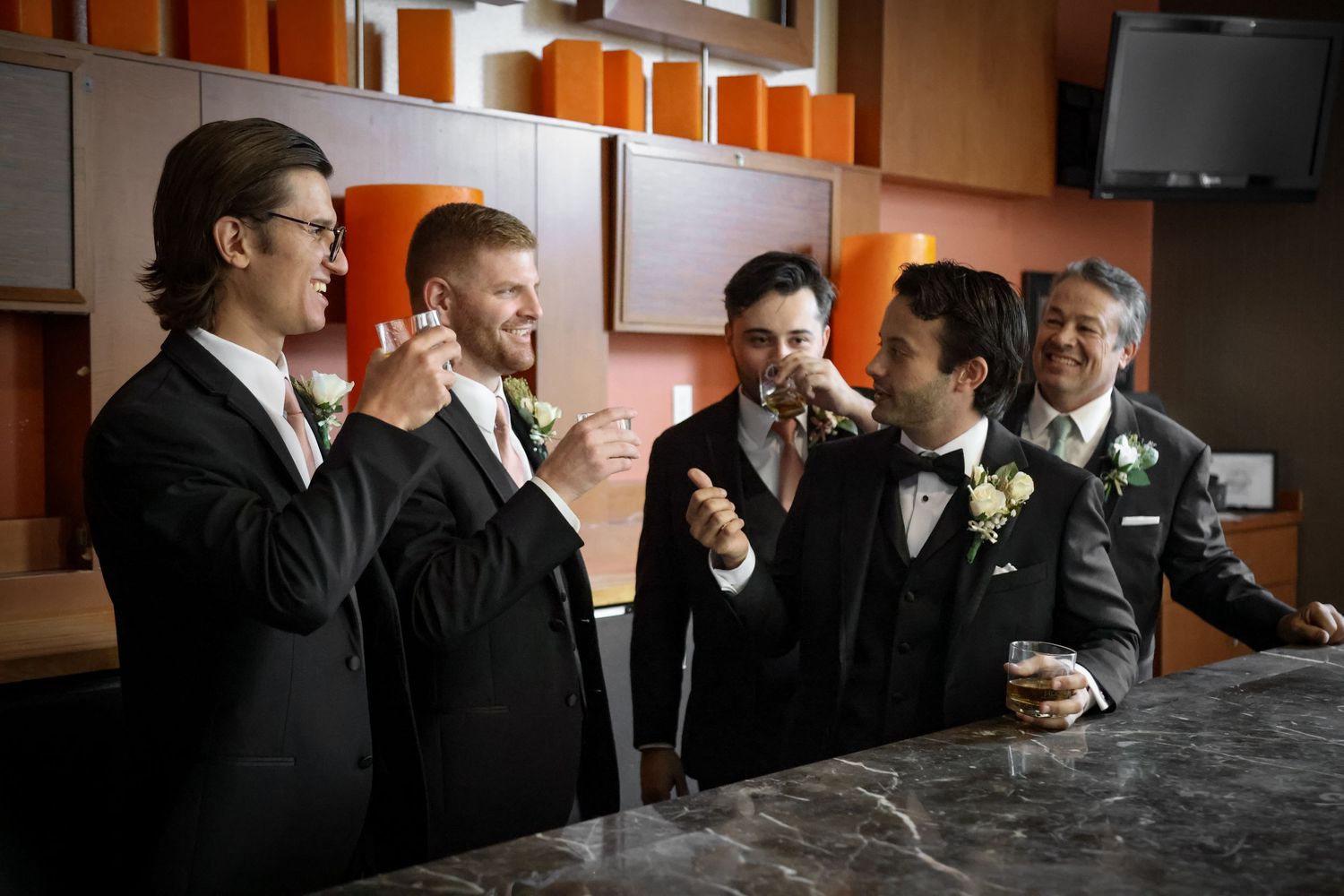 Groomsmen in black tuxedos and bow ties share drinks and laughs at a bar counter during a wedding celebration.