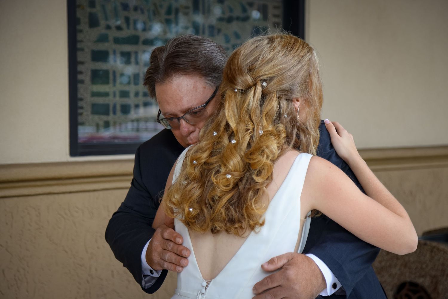 A tender moment at a wedding as two people embrace, with the bride wearing a white dress and having long curled blonde hair.