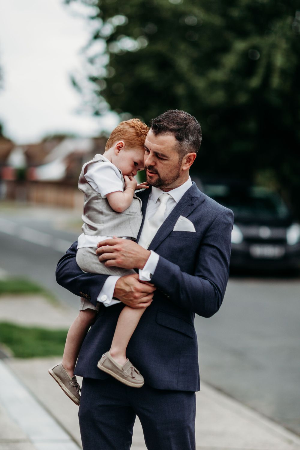 Person in navy suit holding small child outdoors on wedding day.