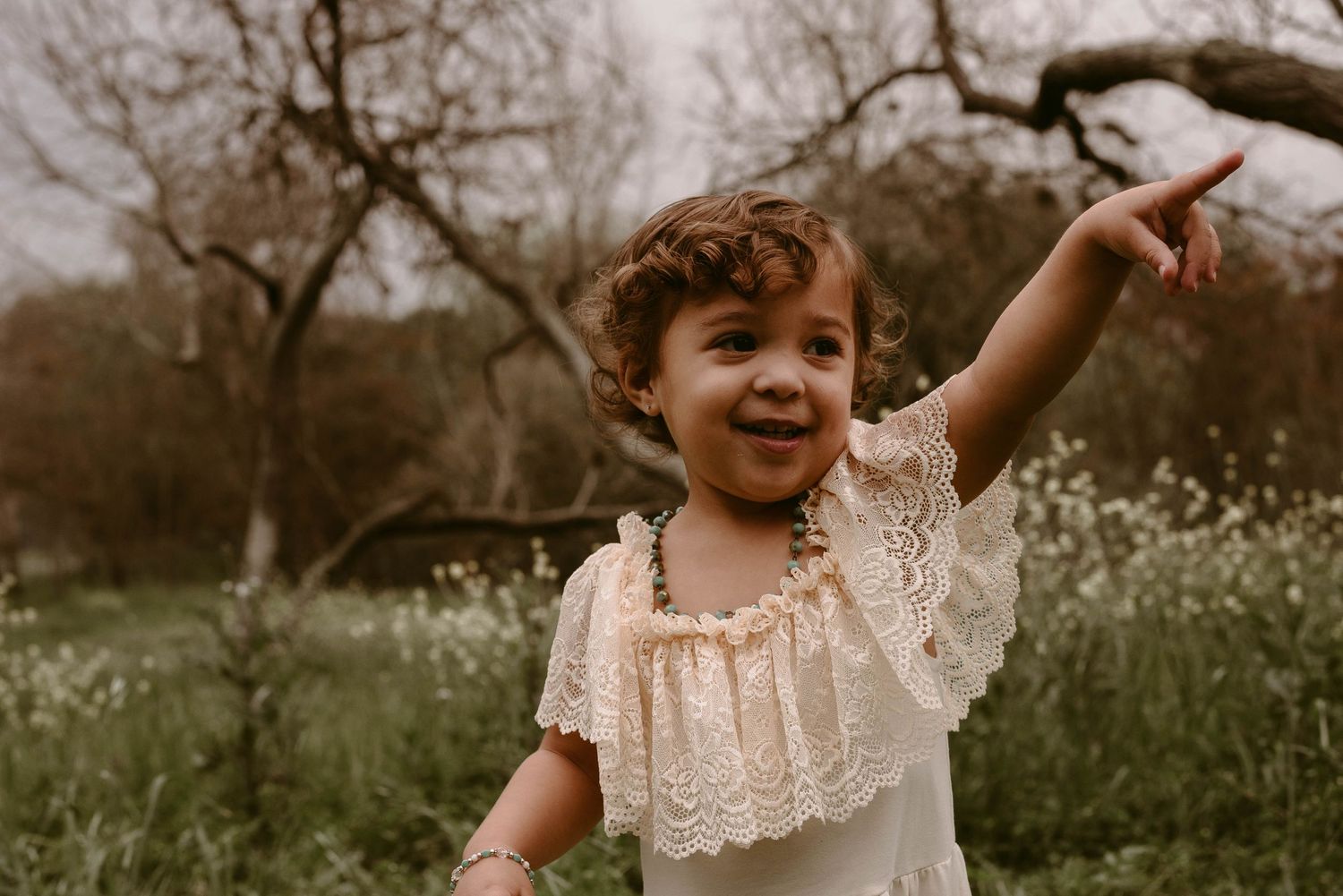 Young child in lace dress points excitedly while playing in a field of flowers.