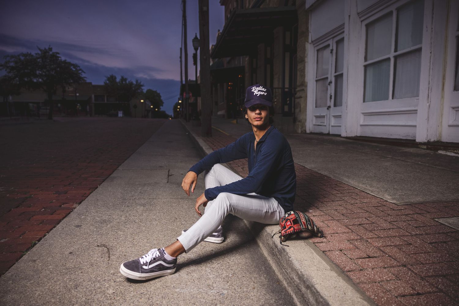 Person sits casually on brick sidewalk at dusk wearing baseball cap and sneakers.