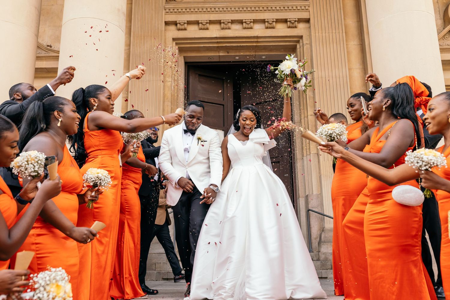 Joyful wedding celebration with bridesmaids in vibrant orange dresses throwing flower petals at church entrance.