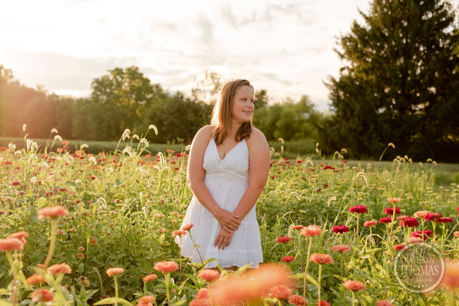 Someone wearing white dress stands among blooming orange zinnia flowers during golden hour.